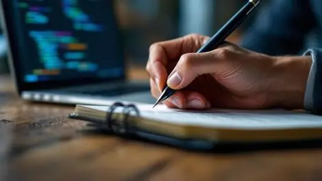 Hand holding pen writing notes paper notebook laptop coding background